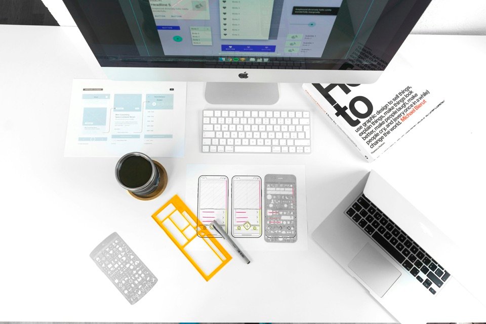 Un bureau vu du dessus : écran, ordinateur portable, croquis d'écrans de téléphone et règle à dessin.